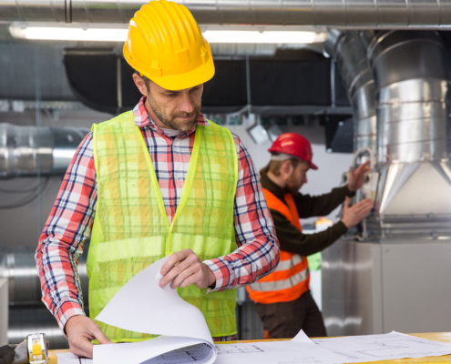 Front view of man looking at blueprints while other man in background inspects HVAC ducts